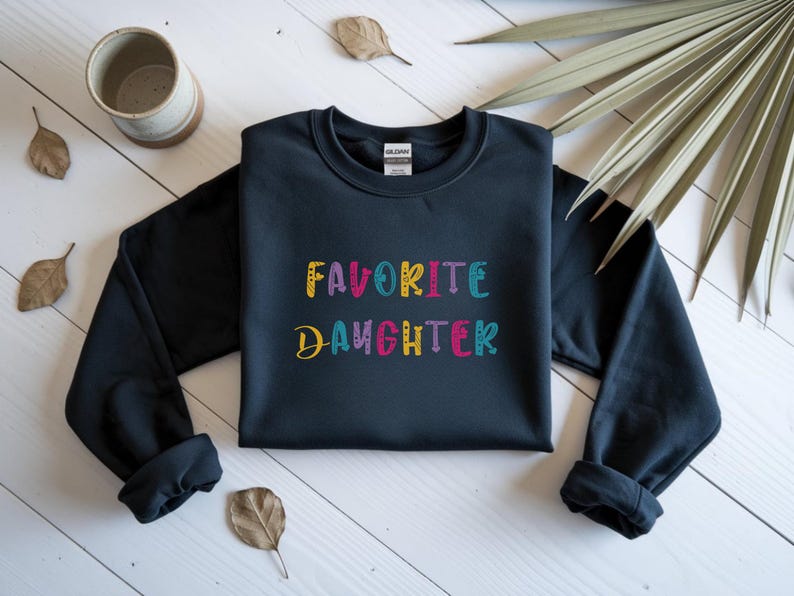 Op de afbeelding: Een marineblauwe sweatshirt met de tekst "FAVORITE DAUGHTER" in kleurrijke, geborduurde letters. De sweatshirt wordt gepresenteerd op een wit houten oppervlak, met decoratieve bladeren en een keramische mok op de achtergrond.