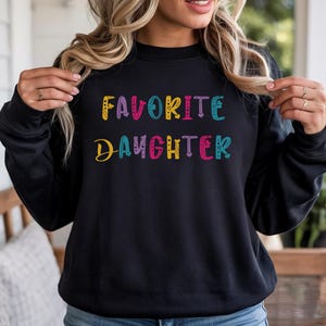 Op de afbeelding: Zwarte sweatshirt met de tekst "FAVORITE DAUGHTER" in kleurrijke, speelse letters. De tekst is in een mix van lettertypen en kleuren, waaronder geel, roze, blauw en paars. Een casual, comfortabele top.