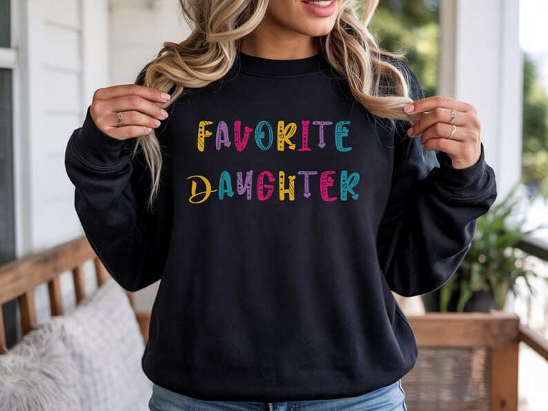 Op de afbeelding: Zwarte sweatshirt met de tekst "FAVORITE DAUGHTER" in kleurrijke, speelse letters. De tekst is in een mix van lettertypen en kleuren, waaronder geel, roze, blauw en paars. Een casual, comfortabele top.