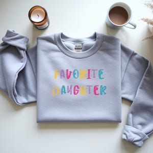 Op de afbeelding: Een lichtgrijs sweatshirt met de tekst "FAVORITE DAUGHTER" in kleurrijke, speelse letters. Een brandende kaars in een bruine glazen pot en een mok koffie zijn zichtbaar op de achtergrond.