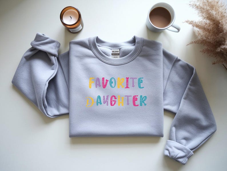 Op de afbeelding: Een lichtgrijs sweatshirt met de tekst "FAVORITE DAUGHTER" in kleurrijke, speelse letters. Een brandende kaars in een bruine glazen pot en een mok koffie zijn zichtbaar op de achtergrond.