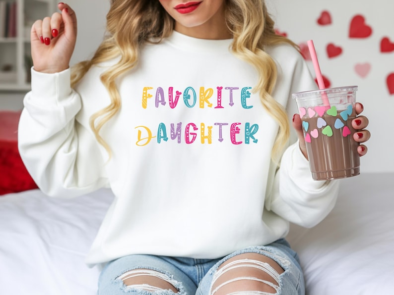 Op de afbeelding: Witte sweatshirt met de tekst "FAVORITE DAUGHTER" in kleurrijke letters. De persoon houdt een doorzichtige plastic beker vast met een roze rietje en een chocoladekleurige drank, versierd met hartvormige stickers.