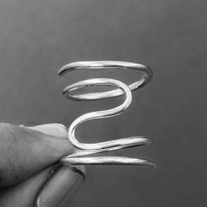May include: A silver-toned, abstract ring with a spiral design. The ring features a series of curved lines that create a unique, sculptural shape. The ring is held by a hand against a dark background.