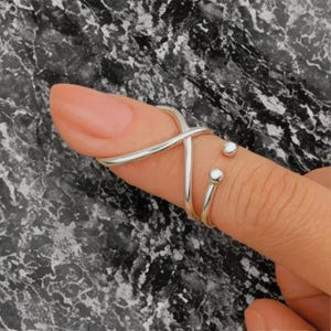May include: A silver-colored finger ring with a unique design. The ring features a crisscross design on the top of the finger and a smaller ring with two small, round accents. The ring is worn on a finger with a light pink nail.