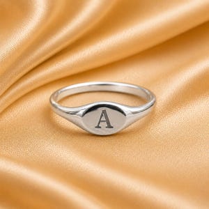 Può includere: Un anello con sigillo d'argento con finitura lucida. L'anello presenta una faccia ovale con la lettera maiuscola "A" incisa al centro. L'anello è posto su uno sfondo di raso dorato.