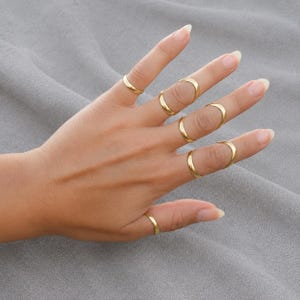 Könnte beinhalten: Eine Hand, die mehrere goldfarbene Ringe zeigt. Die Ringe haben unterschiedliche Designs, darunter schlichte Bänder und offene Kreisformen. Die Ringe werden an mehreren Fingern getragen und präsentieren den Schmuck.