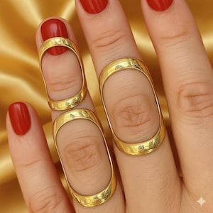 May include: Gold-colored finger rings with an elongated oval design. The rings are worn on the fingers, with the oval shape encompassing the finger. The nails are painted red. The background is a gold-colored fabric.
