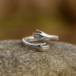 May include: Silver ring with an embracing hands design. The word "Devil" is engraved on the band. The ring is shown on a textured, grey stone surface. The ring's design is detailed.