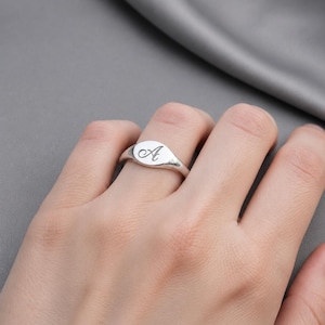 Peut inclure: Une bague chevalière en argent avec une finition polie. La bague présente une face ovale avec la lettre "A" en cursive gravée. La bague est portée au doigt.
