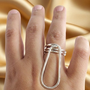 May include: A silver ring with a unique design. The ring features a large, elongated teardrop shape that extends down the finger, connected to a series of smaller bands that wrap around the base of the finger. The ring is made of a shiny, metallic material.