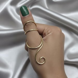 May include: A gold-colored thumb ring with a unique, flowing design. The ring wraps around the thumb with a curved, open design, featuring a spiral at the end. The thumb has a green nail polish.