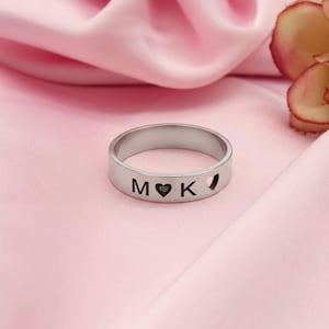 Pode incluir: Um anel prateado com as letras gravadas "M" e "K", separadas por um símbolo de coração. Um pequeno recorte em forma de coração está no lado direito. O anel é exibido em cetim rosa.