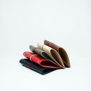 May include: A stack of six leather pen cases in various colors: black, red, green, beige, and brown. Each case is folded and has a contrasting red or white accent. The cases are arranged on a white background.