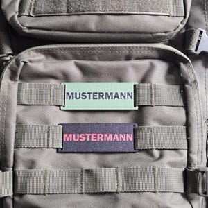 Könnte beinhalten: Nahaufnahme eines taktischen Rucksacks mit zwei Namensschildern. Das obere Schild ist hellgrün mit dem Namen "MUSTERMANN" in Schwarz. Das untere Schild ist dunkelviolett mit dem Namen "MUSTERMANN" in Pink.