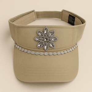 May include: A tan visor with a decorative flower made of clear rhinestones. A silver rhinestone chain is along the brim. The visor has an adjustable strap.