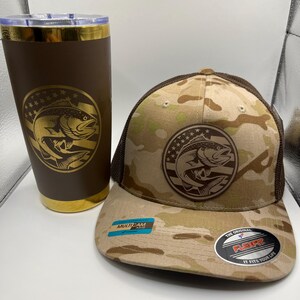 May include: A brown tumbler and a camouflage Flexfit hat. Both items feature a circular emblem with a fish leaping from the water, surrounded by stars. The hat has a brown mesh back and a curved brim. The tumbler has a gold rim.