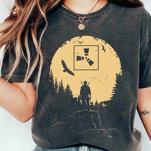 May include: A charcoal gray short-sleeve t-shirt featuring a graphic print of a yellow sun, a silhouette of a person standing on a rocky cliff, a flying bird, and a square logo with three triangles. The shirt has a crew neckline and a relaxed fit.