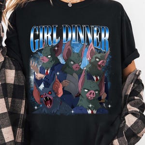 May include: Black t-shirt featuring a graphic of bat-like creatures in suits with the text "GIRL DINNER" in a blue, retro font. The bats have green skin and red eyes. The shirt is worn by a person.