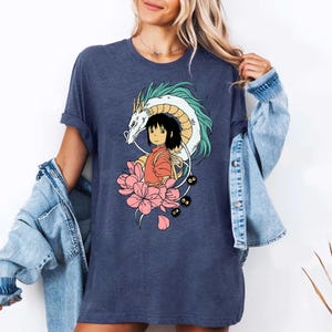 以下が含まれることがあります： ネイビーブルーのTシャツ。アニメ風のカラフルなイラストが特徴です。デザインには、白と水色のアクセントのドラゴン、オレンジ色の服を着た女の子、ピンクの花、小さな黒いキャラクターが含まれています。カジュアルなスタイルです。