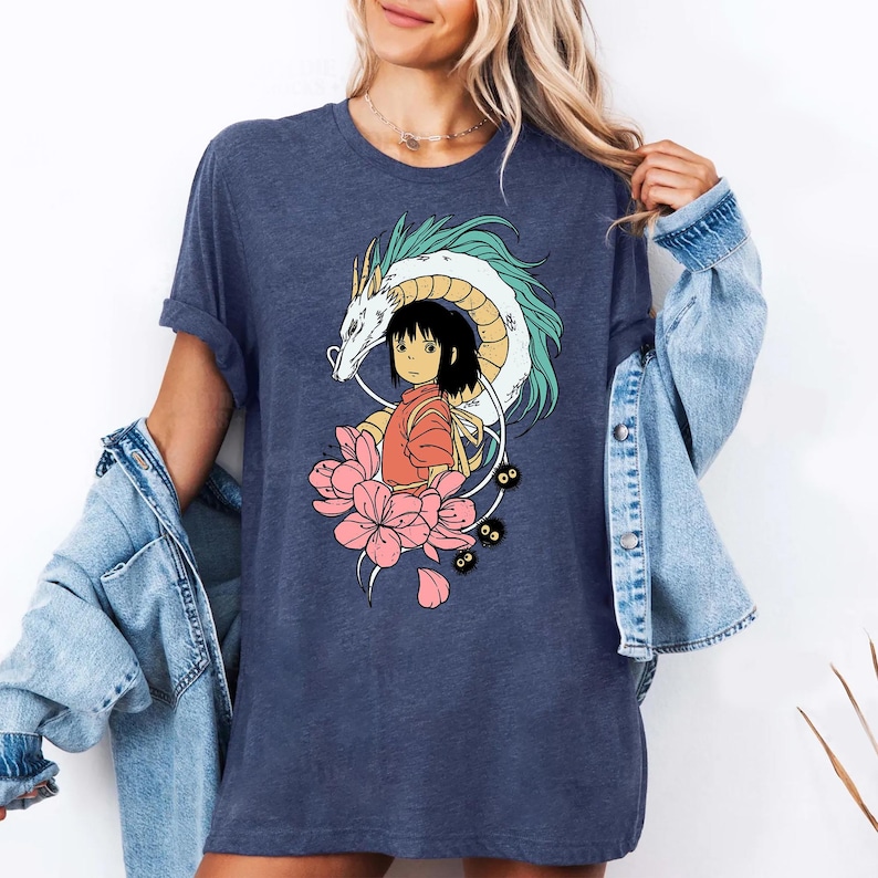 以下が含まれることがあります： ネイビーブルーのTシャツ。アニメ風のカラフルなイラストが特徴です。デザインには、白と水色のアクセントのドラゴン、オレンジ色の服を着た女の子、ピンクの花、小さな黒いキャラクターが含まれています。カジュアルなスタイルです。