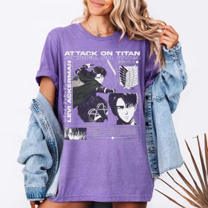 Pu&ograve; includere: Una maglietta viola con grafica della serie anime "Attack on Titan". Il design include immagini di Levi Ackerman e il logo della serie in bianco. La maglietta &egrave; indossata con una giacca di jeans azzurra.