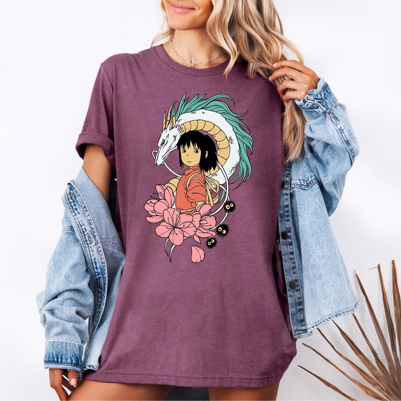 以下が含まれることがあります： アニメ風のカラフルなグラフィックが特徴のマルーンTシャツ。デザインには、ターコイズのアクセントが付いた白いドラゴン、オレンジ色の服を着た少女、ピンクの花、小さな黒いキャラクターが含まれています。Tシャツは半袖です。