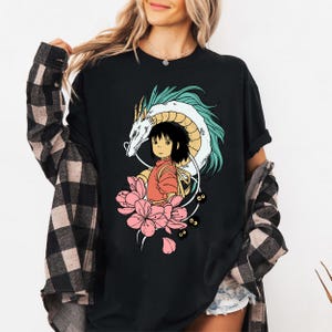 以下が含まれることがあります： 黒いTシャツに、カラフルなアニメ風イラスト。白いドラゴン、赤い服を着た少女、ピンクの花、小さな黒い姿が描かれています。アートワークは黒い背景に配置されています。