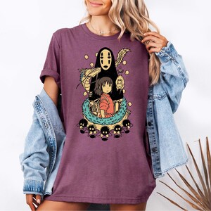 Może przedstawiać: Fioletowa koszulka z kolorową grafiką z filmu animowanego Spirited Away. Projekt przedstawia postacie takie jak No-Face, Chihiro i smoka, otoczone innymi fantastycznymi elementami. Koszulka ma swobodny, codzienny styl.