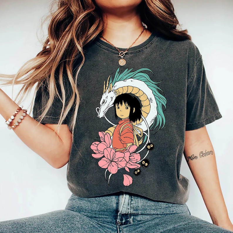 以下が含まれることがあります： アニメスタイルのカラフルなグラフィックが特徴のダークグレーのTシャツ。デザインには、若い女の子、ターコイズアクセントの白いドラゴン、ピンクの蓮の花が含まれています。このシャツは、快適でカジュアルなスタイルです。