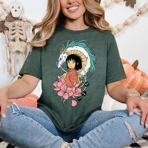 以下が含まれることがあります： 濃い緑色のTシャツ。カラフルなアニメスタイルのグラフィックが特徴です。デザインには、ターコイズのアクセントが付いた白いドラゴン、赤い服を着た少女、ピンクの花、小さな黒い生き物が含まれています。