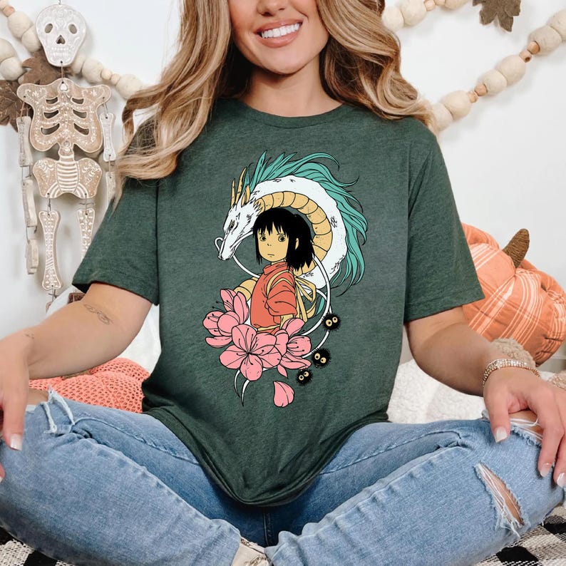 以下が含まれることがあります： 濃い緑色のTシャツ。カラフルなアニメスタイルのグラフィックが特徴です。デザインには、ターコイズのアクセントが付いた白いドラゴン、赤い服を着た少女、ピンクの花、小さな黒い生き物が含まれています。