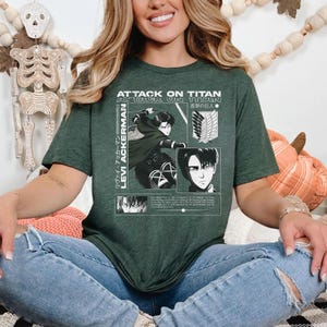 Pu&ograve; includere: Una maglietta verde scuro con la scritta "ATTACK ON TITAN" e "LEVI ACKERMAN" in bianco. La maglietta presenta una grafica di un personaggio della serie anime. La maglietta &egrave; indossata da una persona.