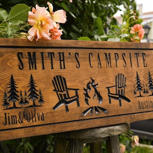 May include: A rectangular wooden sign with a dark brown stain featuring the engraved text SMITH'S CAMPSITE at the top. Below, it displays carved graphics of two pine tree clusters, two Adirondack chairs, and a campfire, with Jim & Oliva and Austin, TX.
