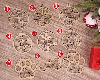 May include: Wooden Christmas ornaments with names like John, Michael, and Connor, along with snowflake and tree designs. Other ornaments include a dog bone and paw print with names. The ornaments are made of light brown wood.