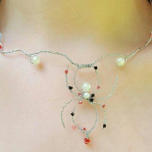 May include: A silver wire necklace featuring a spider pendant. The necklace has a delicate design with red, black, and white beads. The spider pendant has a pearl-like bead at its center.