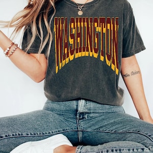 May include: A charcoal grey short-sleeve t-shirt featuring the word WASHINGTON printed in a large, arched, distressed font. The lettering is coloured in a deep burgundy red with a bright yellow outline, creating a bold, layered visual effect.