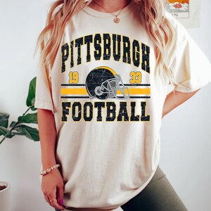 May include: A cream-coloured short-sleeve t-shirt featuring a distressed black and yellow graphic. The design includes the text PITTSBURGH FOOTBALL, the numbers 19 and 33, and a central illustration of a football helmet with a face mask.