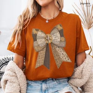 May include: Rust-colored t-shirt featuring a large bow design with a cross and the words "Grace upon grace." The bow is made of various patterned fabrics in shades of brown, beige, and tan. The shirt has short sleeves and a crew neck.