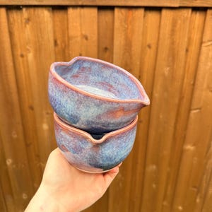 May include: Two stacked ceramic bowls with a rounded, organic shape and a small pouring spout. The glaze features a mottled blend of deep blue, light periwinkle, and warm reddish-brown tones, creating a smooth, glossy finish on the exterior.
