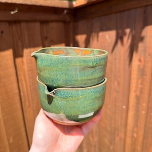May include: Two stacked ceramic bowls with a green speckled glaze and a small pouring spout on each rim. The bowls feature a rounded shape with a textured surface and a visible brown clay base at the bottom of the lower bowl, showing a rustic finish.