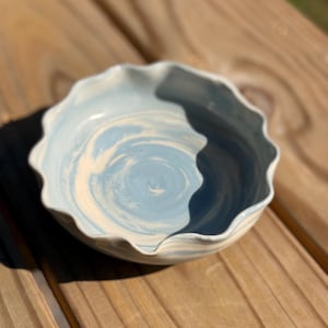 Puede incluir: Un cuenco de cerámica con borde ondulado, con un diseño veteado en azul claro y blanco. El cuenco está sobre una superficie de madera, mostrando su forma única y variaciones de color. Este cuenco hecho a mano es ideal para servir comida o como pieza decorativa.
