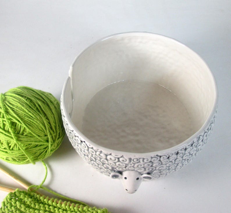 May include: White ceramic yarn bowl shaped like a sheep with black detailing. The bowl has a large opening and a smooth, white interior.