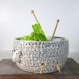 May include: A ceramic yarn bowl shaped like a sheep with a gray and white swirl pattern. The bowl has holes for knitting needles and is filled with green yarn and two wooden knitting needles.