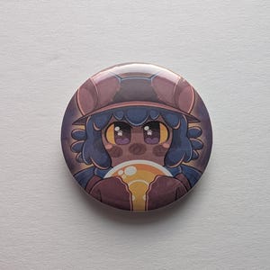 May include: A round button with a cartoon illustration of a character with blue hair, a brown hat, and a brown coat. The character is holding a yellow orb. The button has a glossy finish.