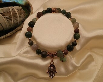 Puede incluir: Pulsera de cuentas con piedras preciosas verdes, marrones y rosas. Tiene un dije de mano de color cobre. La pulsera se exhibe sobre satén crema. Las cuentas se miden en centímetros.