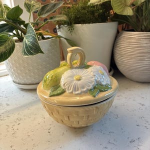 May include: A ceramic trinket box in the shape of a basket, with a lid adorned with a white daisy, a pink fruit, and a green pear. The basket is light yellow and has a woven texture.