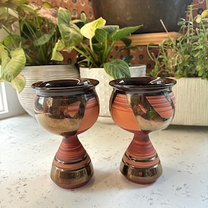 May include: A pair of ceramic goblets featuring a rounded bowl and a tapered stem. The surface displays a combination of matte terracotta-coloured clay with horizontal banding and sections of a dark, metallic, reflective glaze finish.