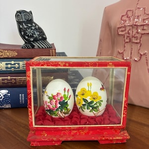 May include: Two decorative eggs with floral designs are displayed inside a red and glass display case. The eggs are white with pink and yellow floral patterns. The case sits on a wooden surface, with a black owl figurine and books in the background.