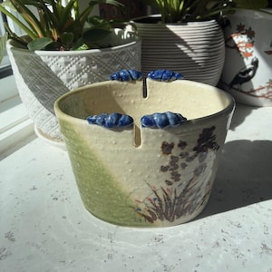 May include: A ceramic yarn bowl with a cream-coloured interior and exterior, featuring a green-glazed side and a hand-painted floral design. The rim has two vertical notches for yarn, accented with small, textured blue decorative ceramic shells.
