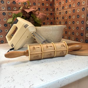 May include: A light beige electric hand mixer, a white ceramic mixing bowl, and a wooden rolling pin with decorative patterns are on a countertop. A small wooden cutting board is also present. The background features a patterned tile backsplash and a potted plant.